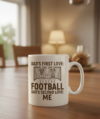 White mug with text on a wooden table in a home setting. This is a Cows Corner mug with a funny football slogan 'Dad’s First Love: Football. Dad’s Second Love: Me'. Cows Corner gifts are perfect for sport-mad fans, these gifts work brilliantly for birthdays, new baby celebrations, Father’s Day, Mother’s Day, Christmas, anniversaries, thank you gifts, end-of-season team awards, graduations, retirements, and just-because moments when you want to raise a smile. 

