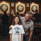 Man and young girl standing together in a pub with dartboards in the background.  The girl is wearing a Cows Corner t-shirt with the funny darts slogan 'I Love My Dad… Even If He Can’t Hit A Double'. Cows Corner gifts are perfect for sport-mad fans, these gifts work brilliantly for birthdays, new baby celebrations, Father’s Day, Mother’s Day, Christmas, anniversaries, thank you gifts, end-of-season team awards, graduations, retirements, and just-because moments when you want to raise a smile. 
