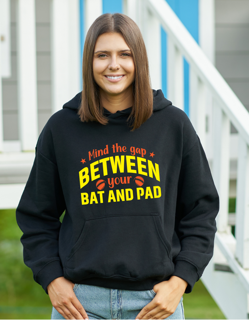 A lady wearing a black hoodie with Mind the Gap Between Your Bat and Pad on the front. A Funny Cricket Hoodie, Premium Fabric, Multiple Adult Sizes