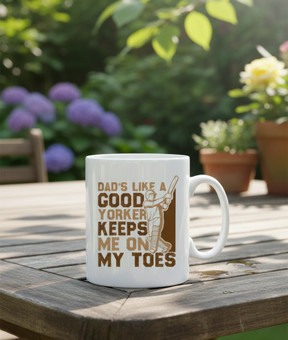 White mug with text and graphic on a wooden table outdoors. This is a Cows Corner slogan with a funny cricket slogan 'Dads Like A Good Yorker, Keeps Me On My Toes'. Cows Corner gifts are perfect for sport-mad fans, these gifts work brilliantly for birthdays, new baby celebrations, Father’s Day, Mother’s Day, Christmas, anniversaries, thank you gifts, end-of-season team awards, graduations, retirements, and just-because moments when you want to raise a smile. 
